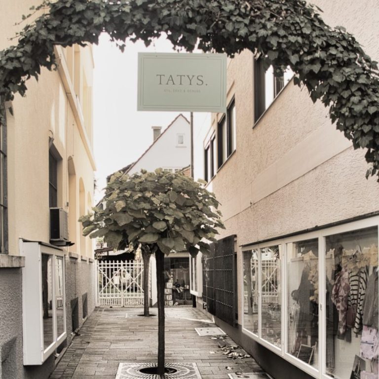 Blick auf einen schmalen Durchgang zu TATYS. mit einer begrünten Überdachung.